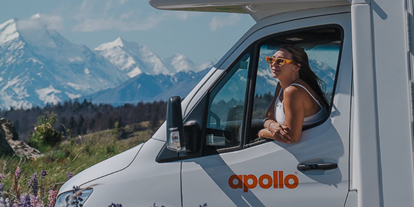 An Apollo campervan parked in a meadow with snow-capped mountains behind