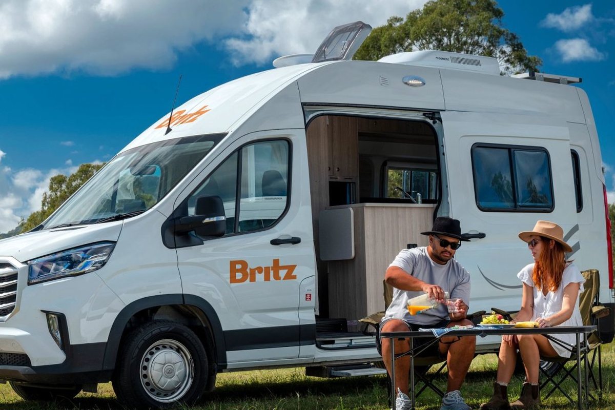 A Britz campervan with the side door open and a couple having breakfast at a table outside