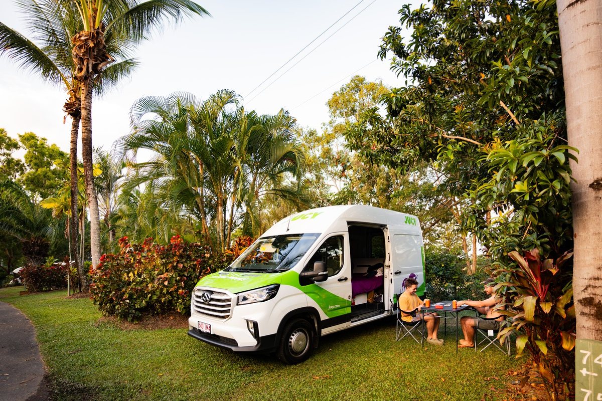 A JUCY campervan parked on grass at a tropical campsite with travellers sitting outside