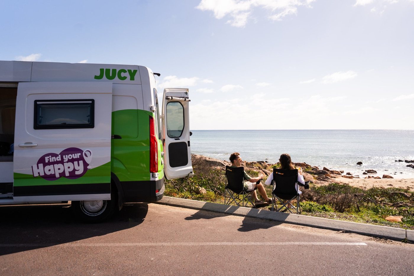 A JUCY campervan parked by the coast with two travellers looking out at the sea