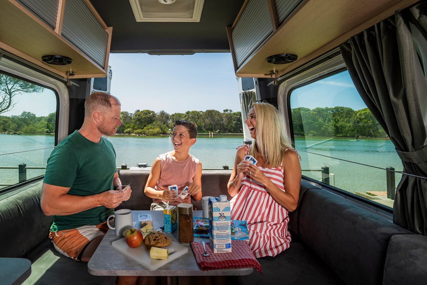 A family having breakfast at the table inside a maui motorhome, water outside the window