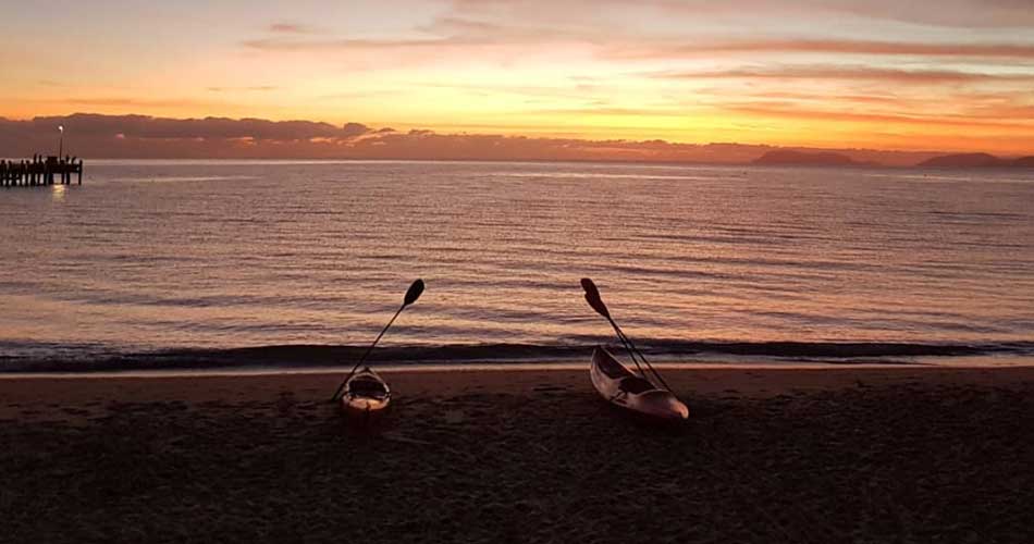 Sunrise over the water at a northern beach near Cairns with kayaks on the sand and a jetty in silhouette